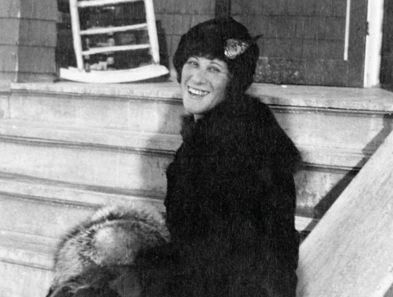 Woman in hat and coat turned sideways smiles on steps in black-and-white photo