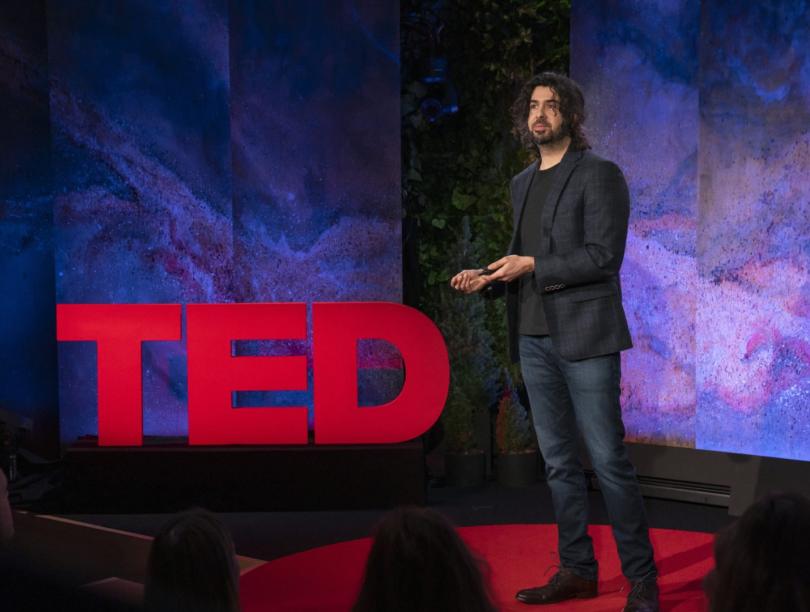 Speaker Dr. Azim Shariff stands on stage before audience in front of a TED sign
