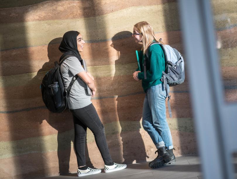 Two students with backpacks talk in front of wall