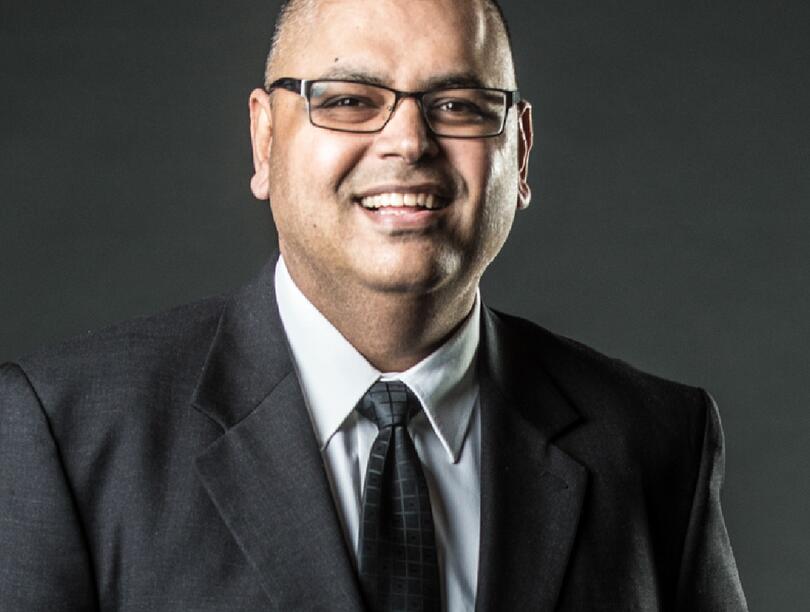 A headshot of Alex Sangha wearing black rectangular glasses and a black suit.