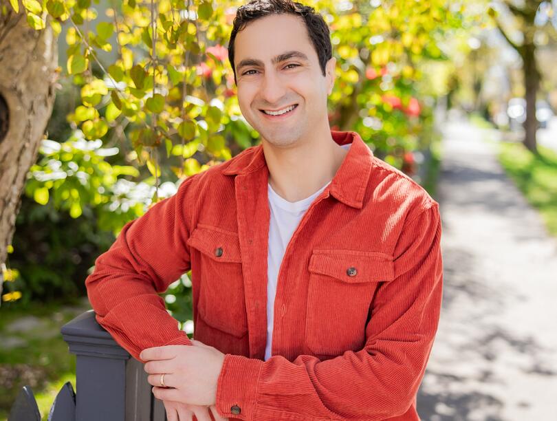 A photo of Saba Farmand standing on a greenery-lined sidewalk, wearing an orange collared jacket.