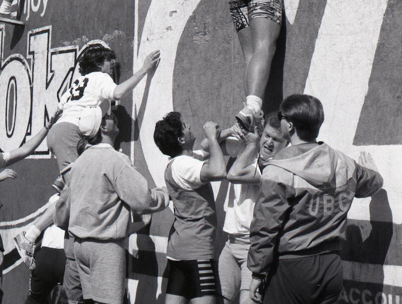 A black and white photo of students pushing each other up a wall.
