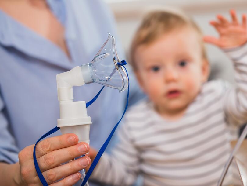 A baby sits in the lap of a nurse who is holding an oxygen mask