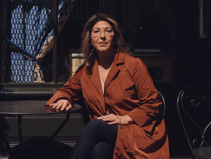 A photo of Naomi Klein seated at a metal table against a dark background, wearing glasses and a russet overcoat.