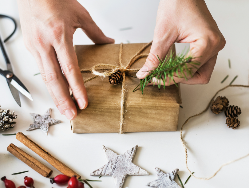 A pair of hands tucks a sprig of spruce into the string wrapped around brown paper package with holiday wrapping materials surrounding it