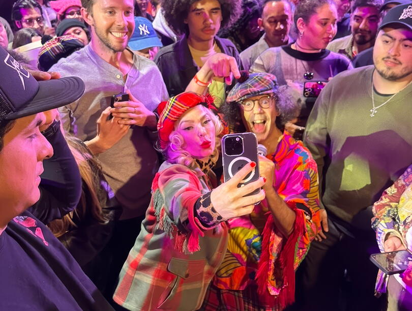 A fan poses with Nardwuar for a selfie, with people milling in the background