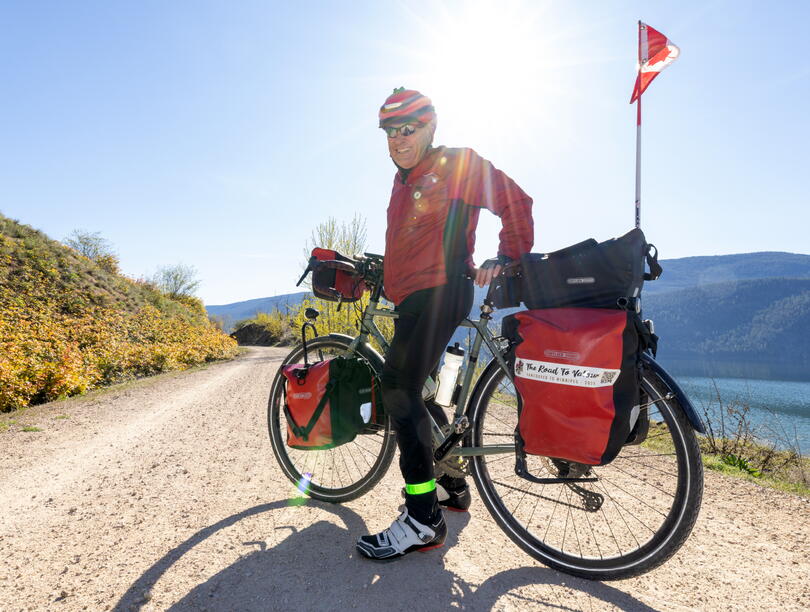 A photo of Garry McCracken standing on a dirt road next to his bike, which has a pannier labelled 'The Road To Valor.' In the background are mountains, water, and a Canadian flag.