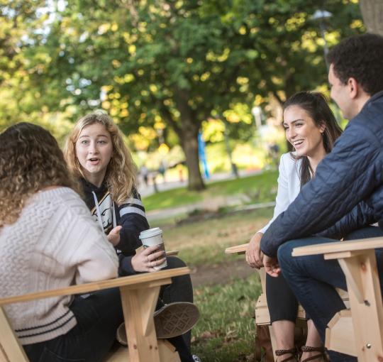Four students sitting on chairs outside, with one student holding a coffee cup