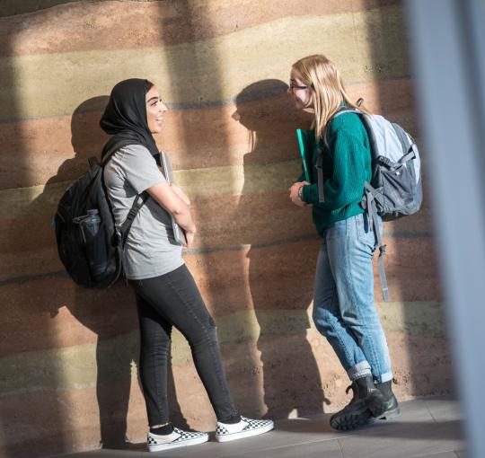 Two students with backpacks talk in front of wall
