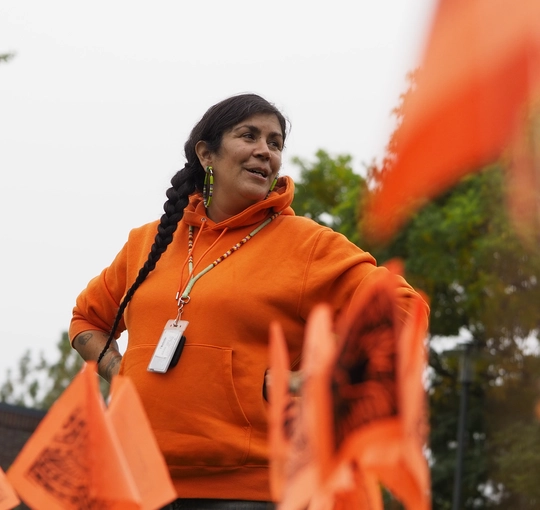 Indigenous woman dressed in orange stands behind small orange flags