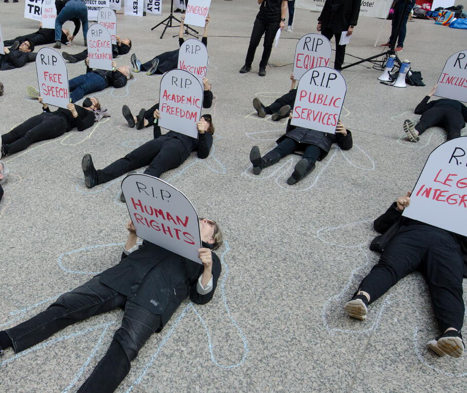 People dressed in black lying on the ground holding tombstone-shaped signs with "RIP" messages related to democracy