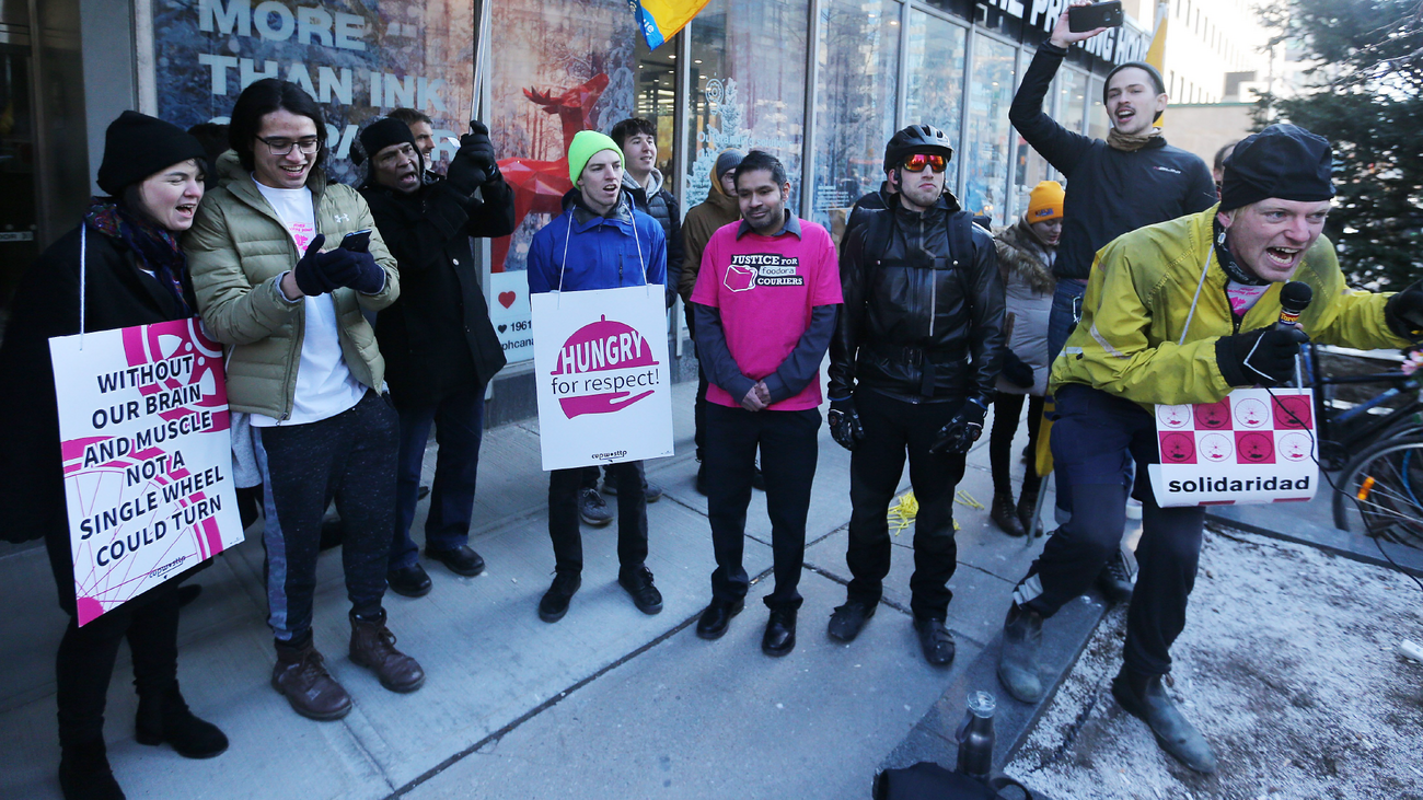 A number of Foodora workers passionately protest on a sidewalk outside of a storefront.