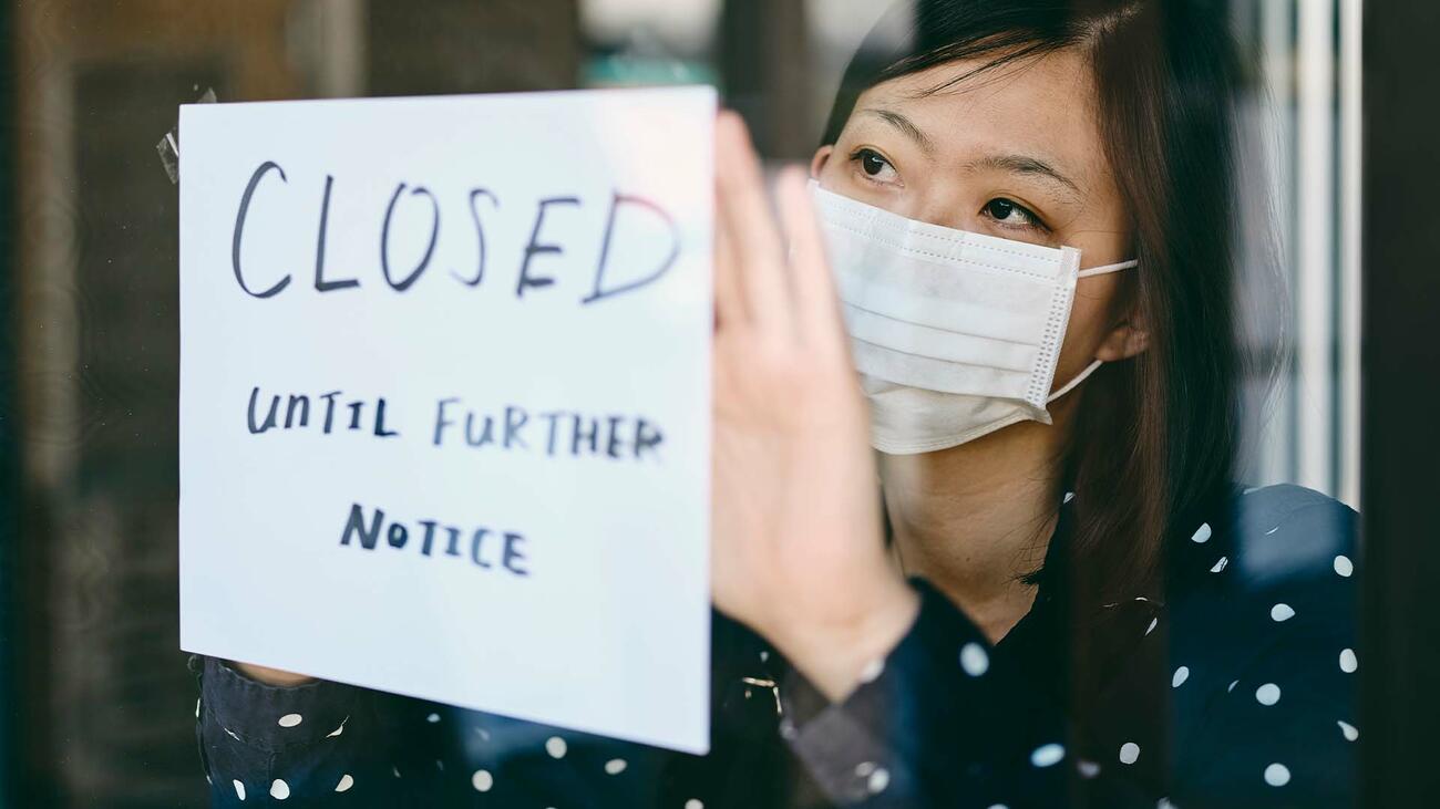 A masked woman puts up a closed sign on a shop.