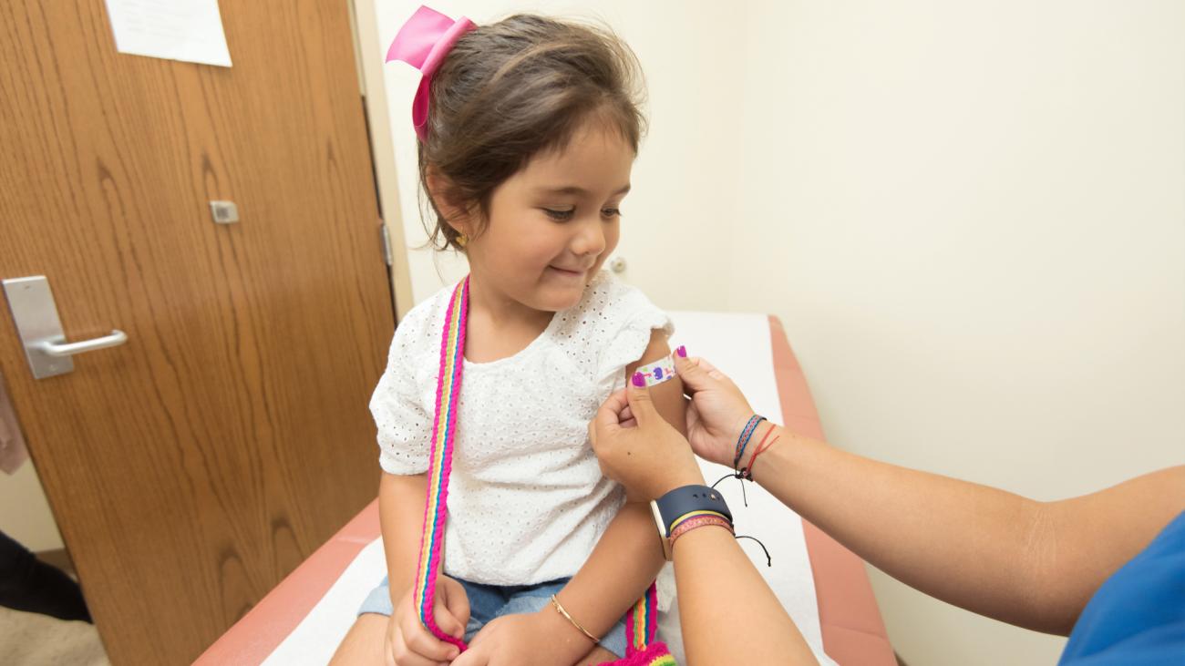Girl sits in a clinic watching healthcare worker apply band-aid to her shoulder