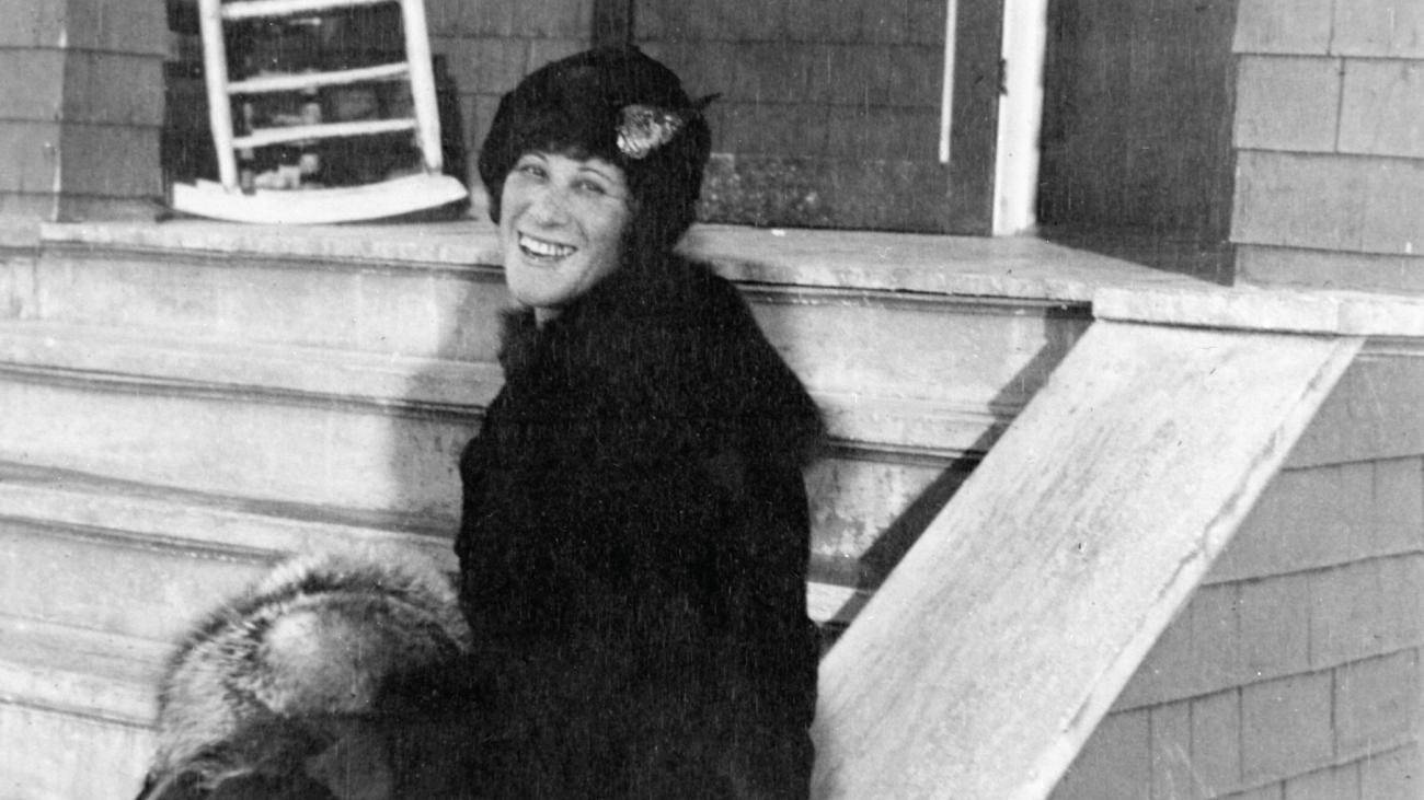 Woman in hat and coat turned sideways smiles on steps in black-and-white photo
