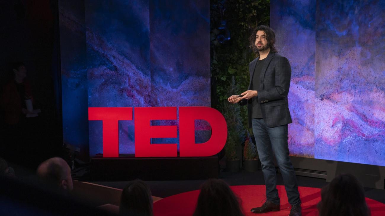 Speaker Dr. Azim Shariff stands on stage before audience in front of a TED sign
