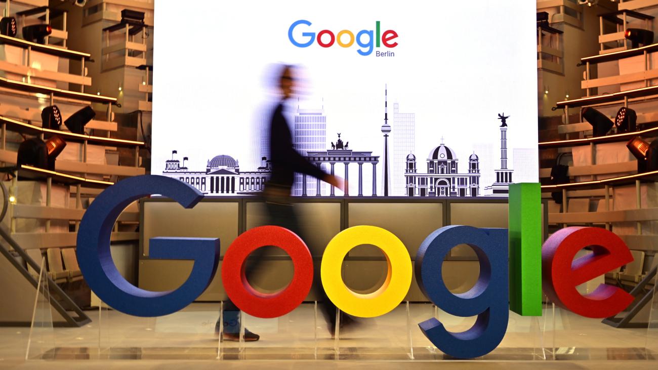 Man walks in front of screen and behind Google logo 