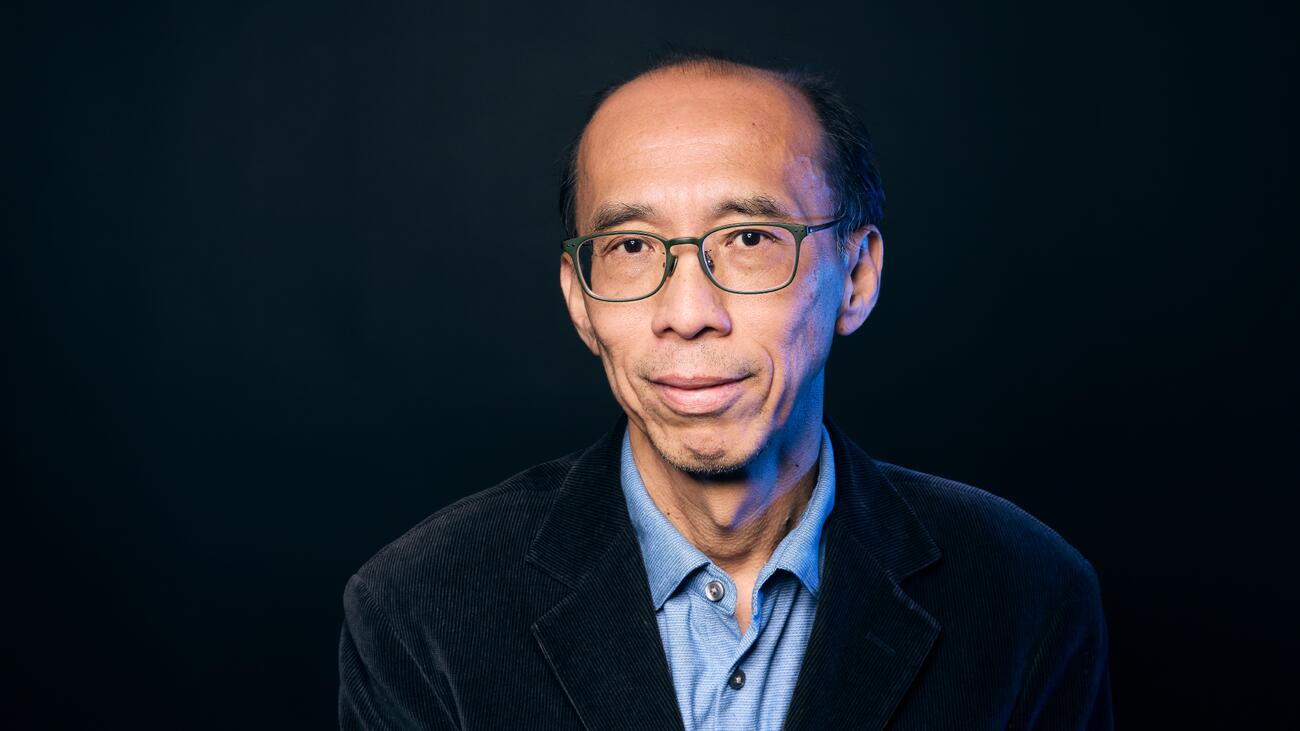 A headshot of Dr. Raymond Ng with dark green glasses, short black hair, and a blue collared shirt against a black backgroun.