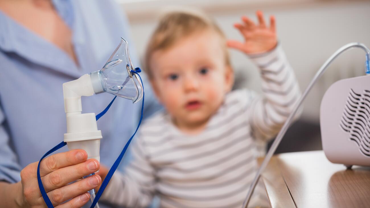 A baby sits in the lap of a nurse who is holding an oxygen mask