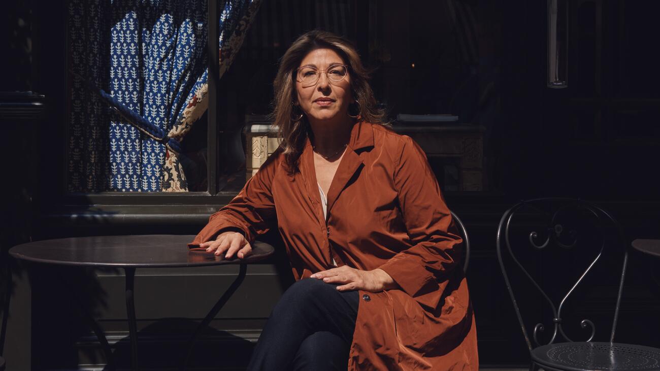 A photo of Naomi Klein seated at a metal table against a dark background, wearing glasses and a russet overcoat.