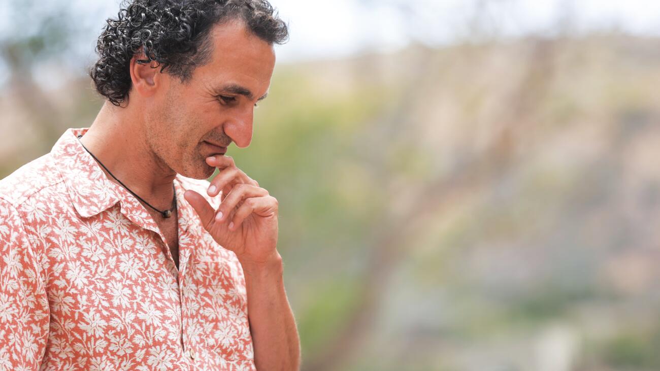 Simon Donner in a pink collared shirt, touching his chin with his finger, against a blurred grassy background.