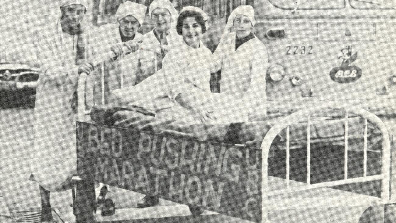  Four people standing around a person in a bed. Attached to the bed is a sign that reads: "UBC Bed Pushing Marathon"