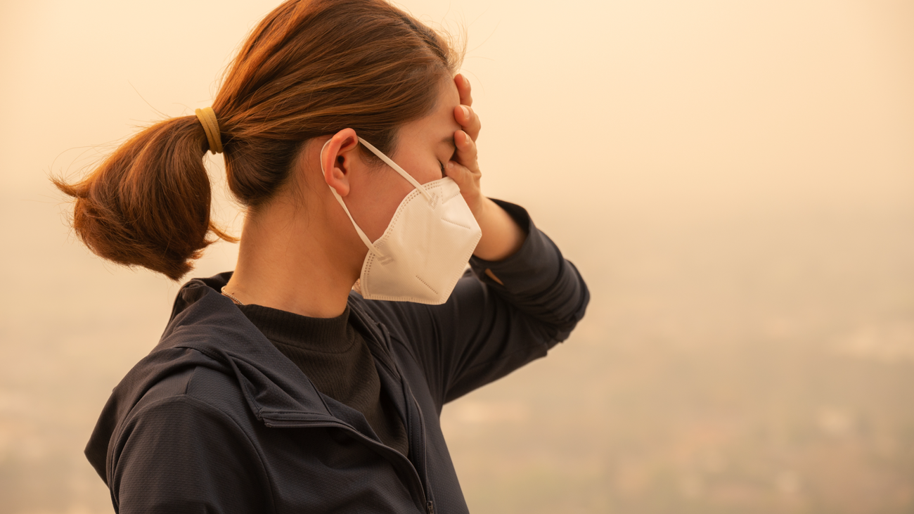 A person in a mask standing in hazy air holds a hand to their forehead