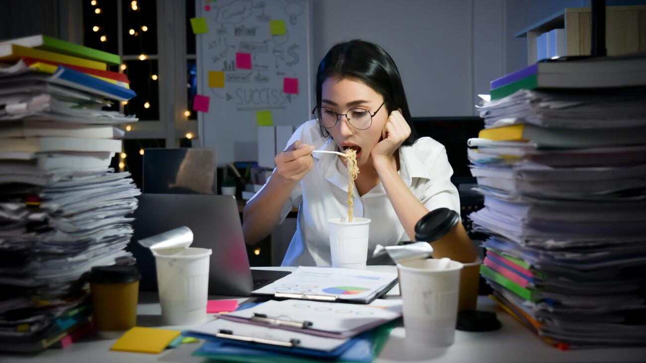Female student eating ramen in front of a laptop and surrounded by piles of schoolwork