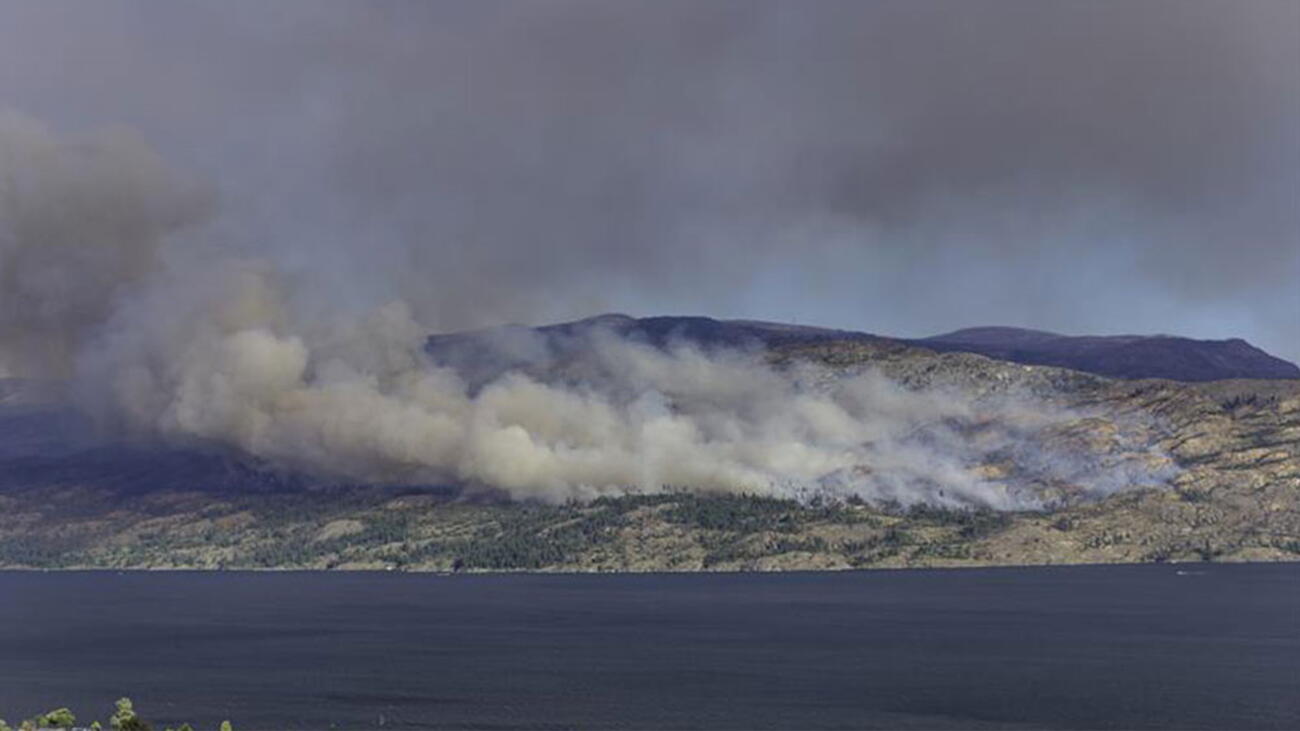 Smoke billowing from a wildfire with water in the foreground and mountains in the background