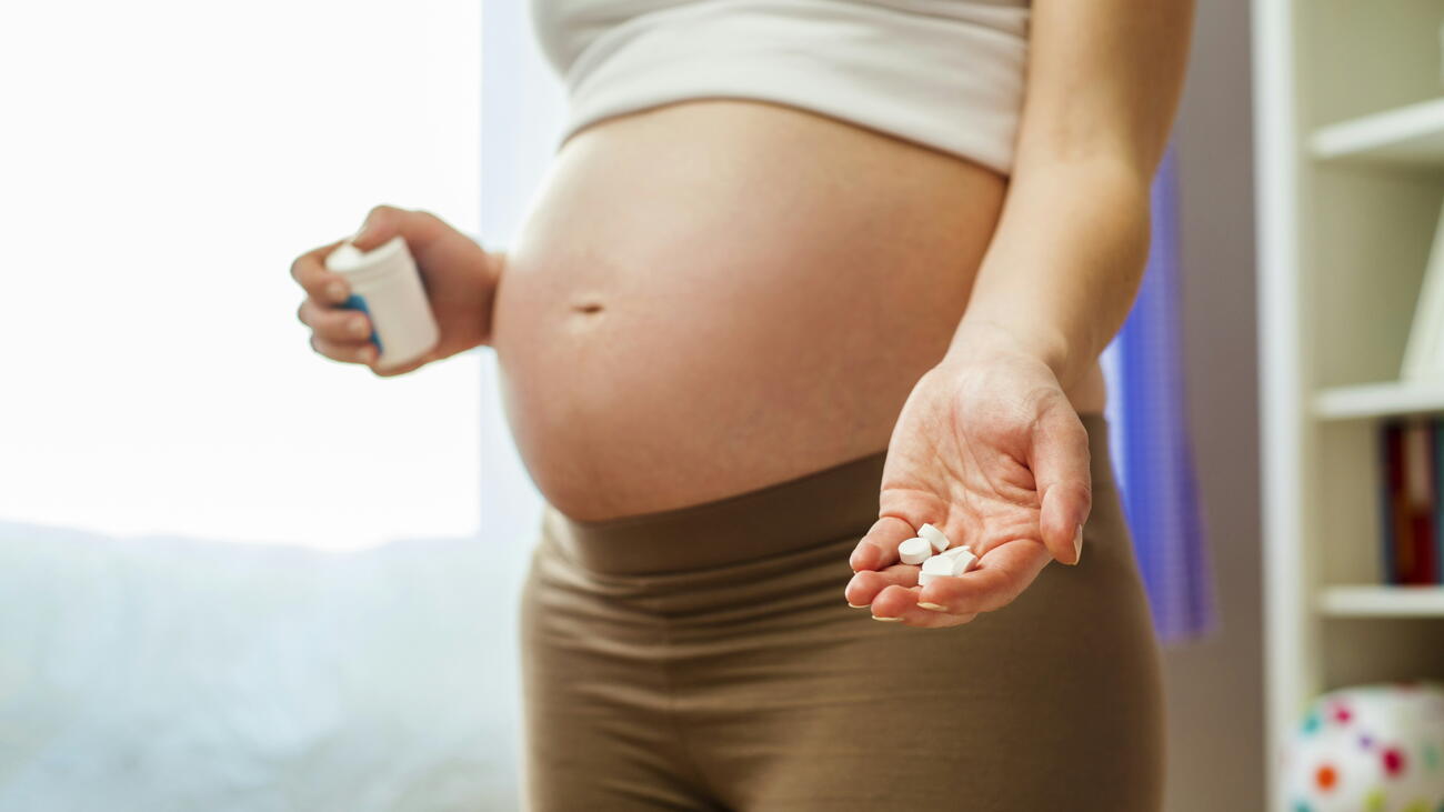 A woman with a pregnant belly, holding a medicine box in one hand and medication pills in the palm of her other hand.
