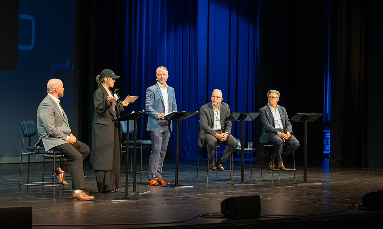 Four men and one woman sit and stand on stage