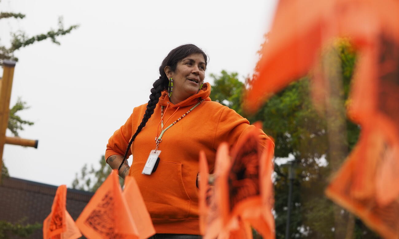 Indigenous woman dressed in orange stands behind small orange flags