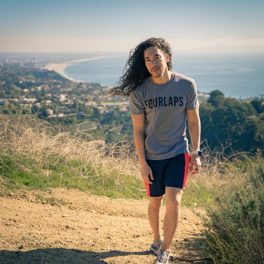 Full-body shot of Cole Walliser, walking on a grassy trail at a high altitude in Pacific Palisades, with the beach behind him
