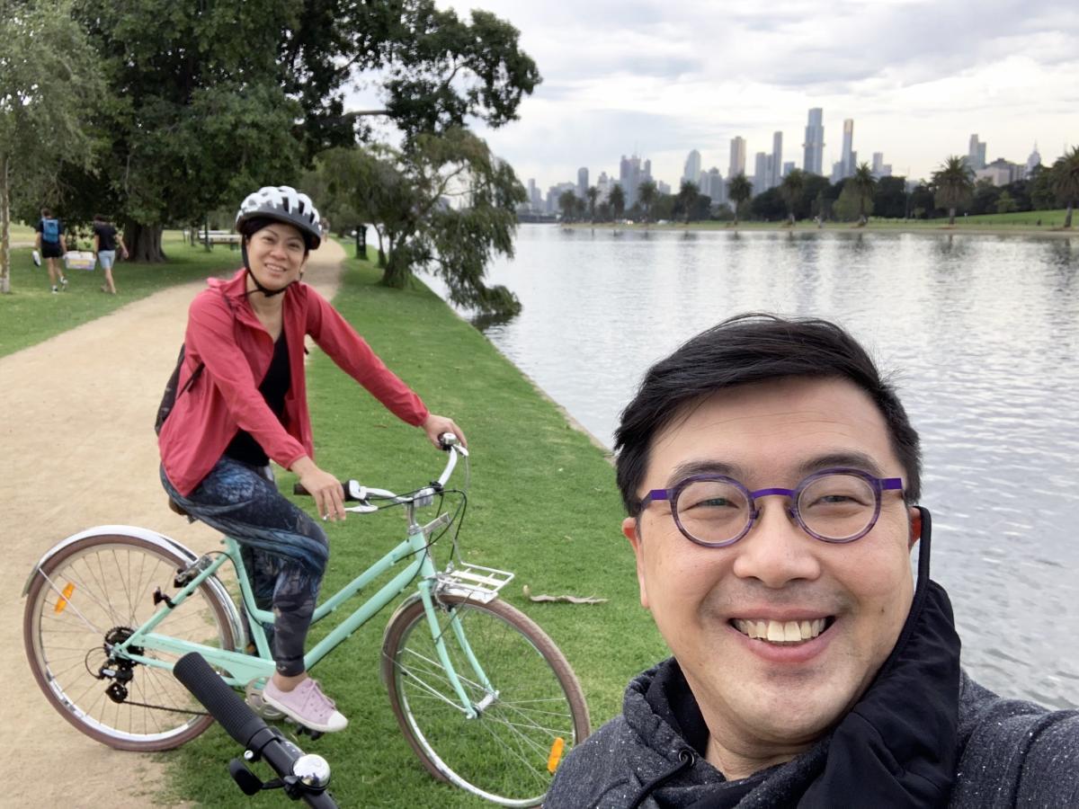 Dave Lim in the foreground with a woman on a bike in the background and a view of water and greenery
