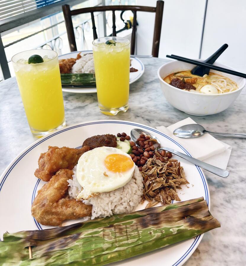 A table with two plate of nasi lemak, a bowl of laksa, and two glasses of calamansi juice