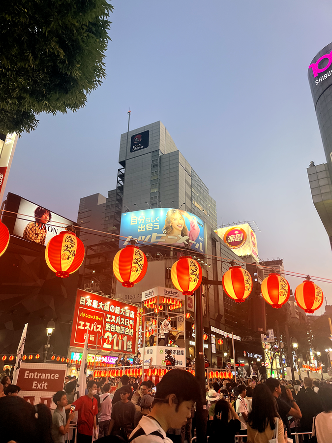People milling on a well-lit street at dusk, with dangling lanterns in the foreground and skyscrapers in the background