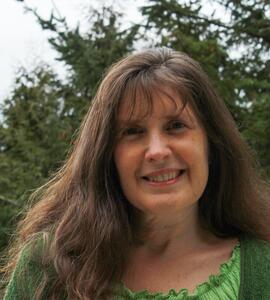 A headshot of Phyllis Dyson, who has long brown hair and is wearing a green shirt and cardigan against a backdrop of trees.
