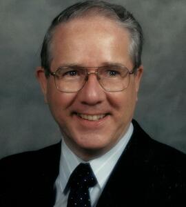 A headshot of Byron Heal with short black and white hair, glasses, and a tuxedo.