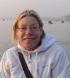 A portrait of Elizabeth Jean Schiller with short blond hair, glasses, and a white jacket, with the ocean behind her.