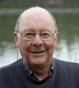 A headshot of Rodney Norman Palmer wearing a navy blue shirt and spectacles in front of a body of water. 