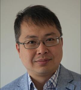 A headshot of Alan Poo wearing glasses and a pale blue suit with a chequered collared shirt.