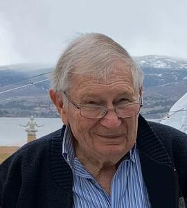 Douglas Arnold Corbishley with white-grey hair and glasses, standing in front of clouded mountains.