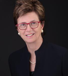 Headshot of Linda Rabeneck with short brown hair and red glasses against a black background