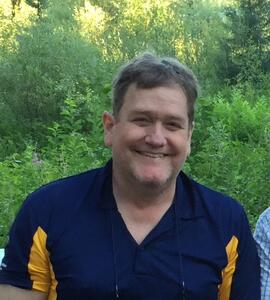 A photo of Bruce Eaket wearing a blue and yellow collared shirt in front of greenery.