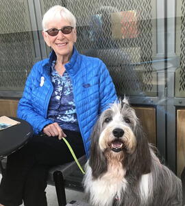 A photo of Patricia Abbott with white hair, dark tinted glasses, and a blue puffer, sitting next to a large black-and-white dog.