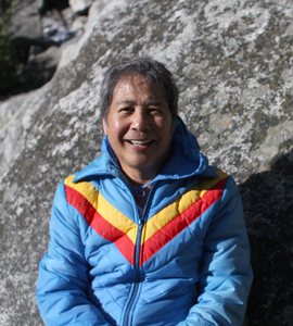 A photo of Chin Choon Khor wearing a light blue puffer with yellow and red stripes in a V-shape. Chin Choon Khor smiles in front of a rocky background.