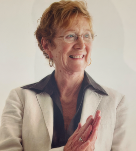 A photo of Julia Levy smiling and looking off-camera, while wearing a white suit jacket and holding her hands together