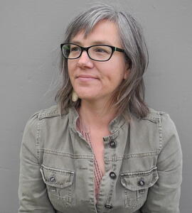 A portrait of Heather Ramsay wearing a grey jean jacket with black glasses and grey-brown shoulder length hair