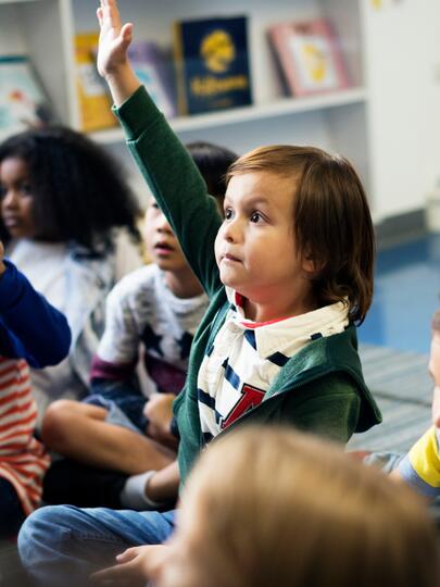Children sitting on floor in classroom, with some holding their hands up