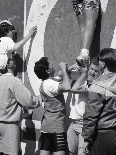 A black and white photo of students pushing each other up a wall.